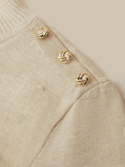 Close-up of a cream-colored knit sweater featuring three decorative gold knotted buttons along the shoulder seam.