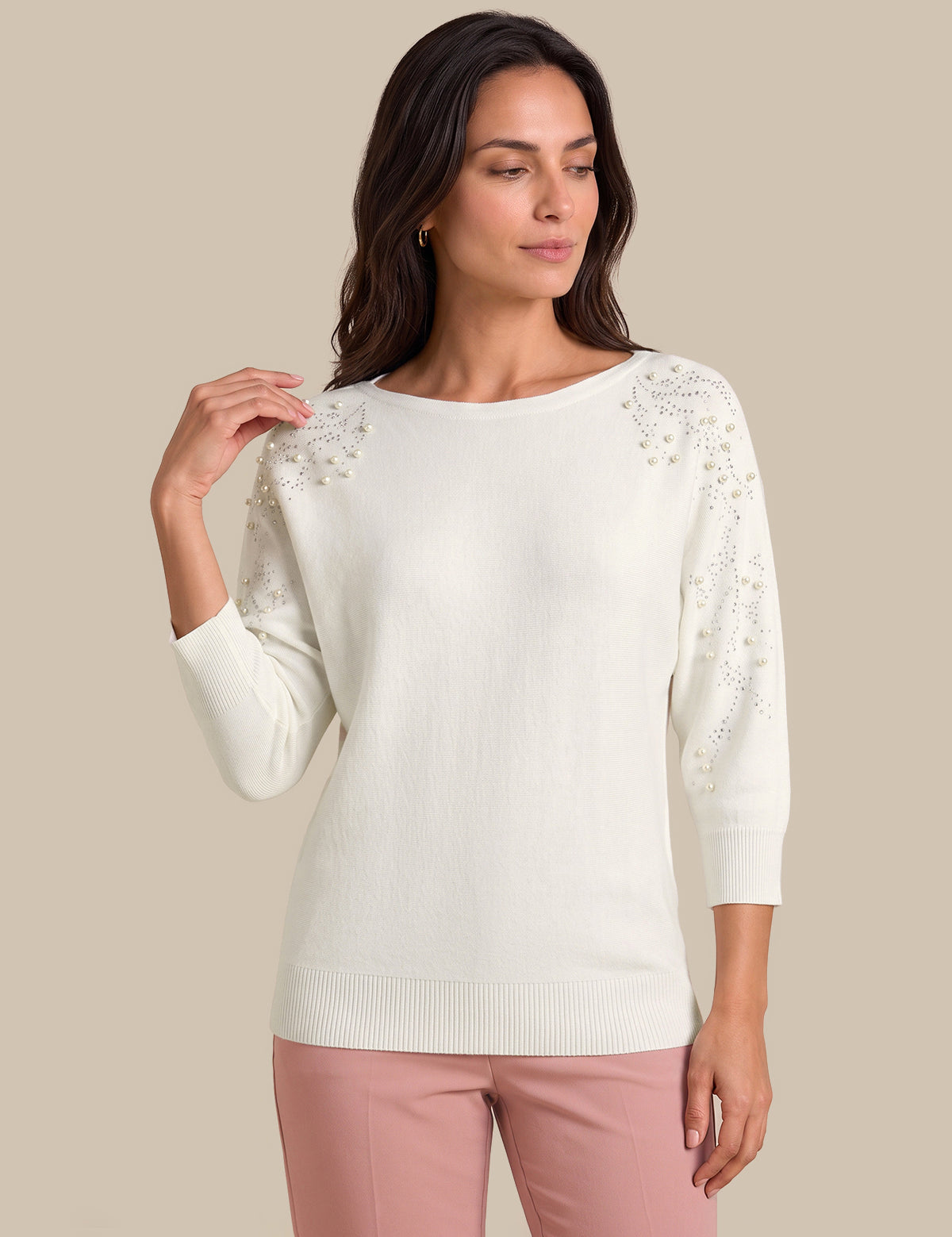 A woman modeling a cream-colored knit sweater with intricate floral lace embroidery on the shoulders and sleeves, paired with dusty pink pants, against a solid beige background.
