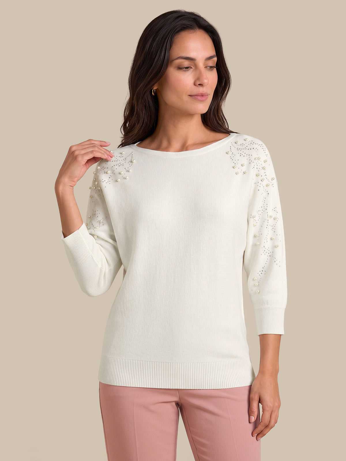 A woman modeling a cream-colored knit sweater with intricate floral lace embroidery on the shoulders and sleeves, paired with dusty pink pants, against a solid beige background.