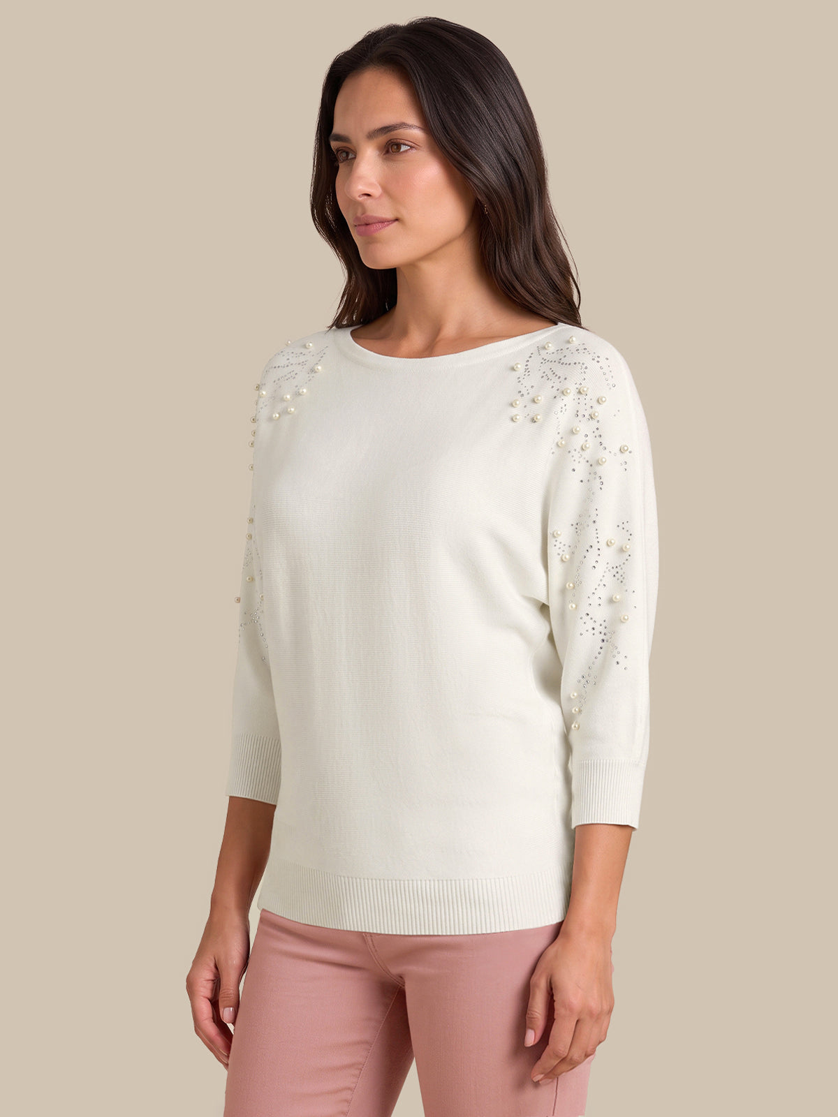 A woman wearing a cream-colored three-quarter sleeve knit sweater with delicate floral embroidery on the shoulders and sleeves, paired with dusty pink pants, set against a solid beige background.