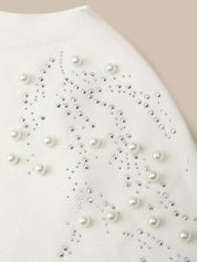 Close-up of a white knit garment decorated with scattered faux pearls and silver rhinestones on a beige background.