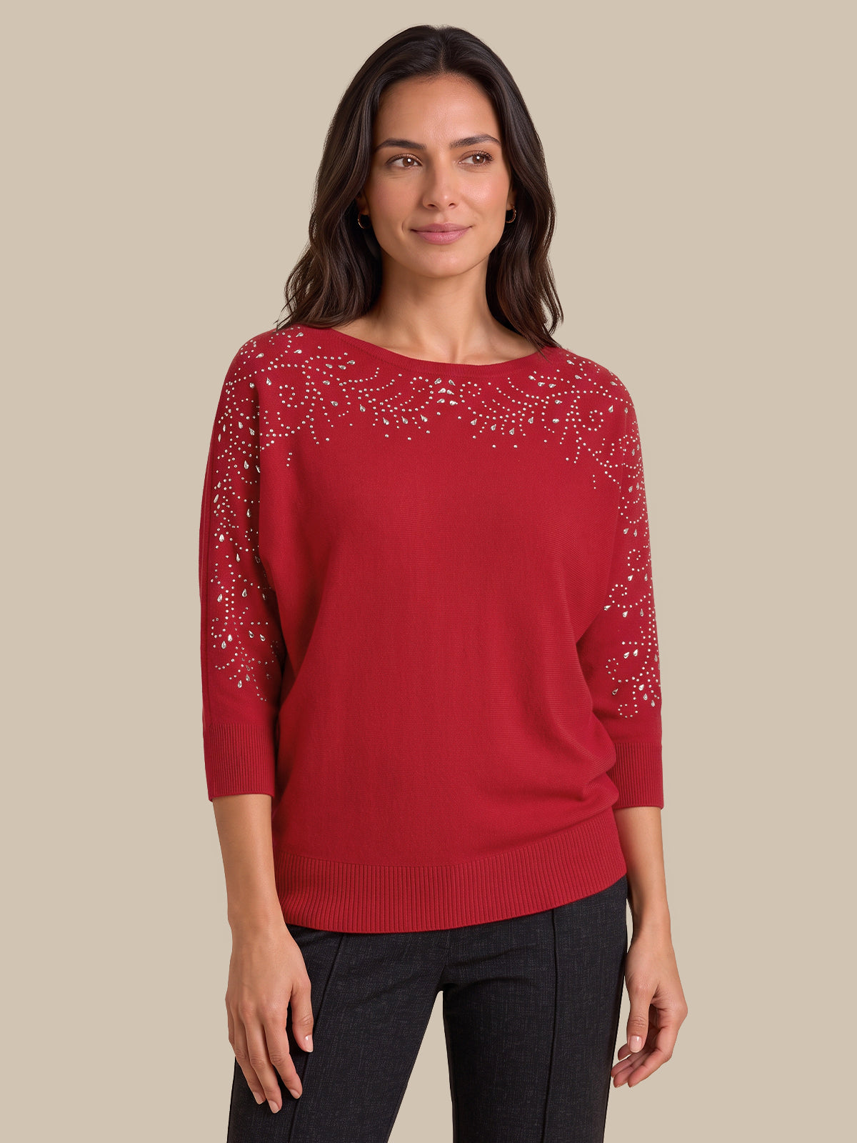 A woman wearing a red knit sweater with silver rhinestone embellishments across the chest and upper sleeves, paired with black pants, against a solid beige background.