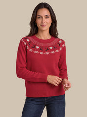 A woman wearing a red crewneck sweater with a decorative yoke pattern of white and black fair isle motifs, paired with dark blue jeans, against a solid beige background.