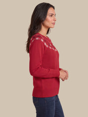 A woman in side profile wearing a red knit sweater with white embroidered detailing across the shoulders, paired with dark blue jeans, against a solid beige background.