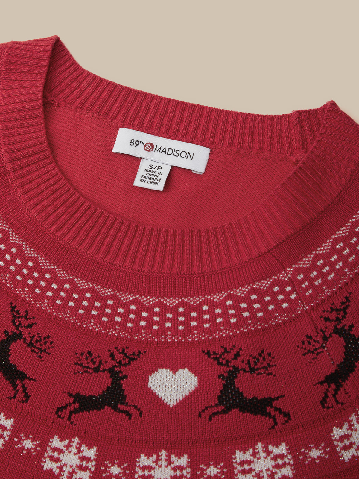 Close-up of a red knit sweater with a white and black holiday pattern featuring reindeer, a white heart, and snowflakes, with a visible 89FIRE MADSON neck label.