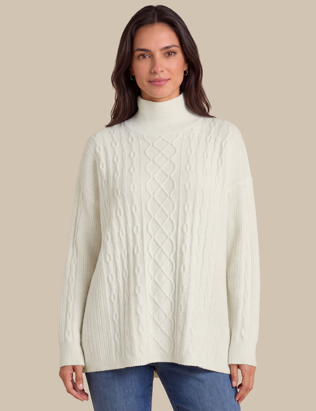 A woman wearing a cream-colored cable-knit mock neck sweater and blue jeans, standing against a solid beige background.