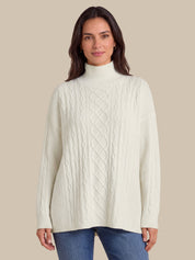 A woman wearing a cream-colored cable-knit mock neck sweater and blue jeans, standing against a solid beige background.