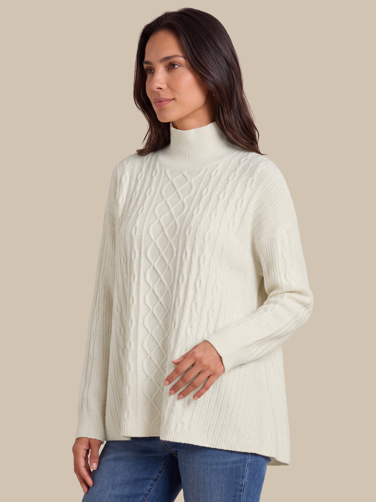 A woman wearing a cream-colored cable-knit mock neck sweater and blue jeans, standing against a solid beige background.