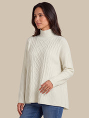 A woman wearing a cream-colored cable-knit mock neck sweater and blue jeans, standing against a solid beige background.