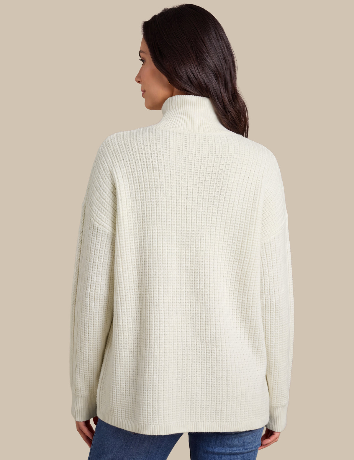 A woman shown from the back wearing a cream-colored, textured waffle-knit mock neck sweater and blue jeans, set against a solid beige background.