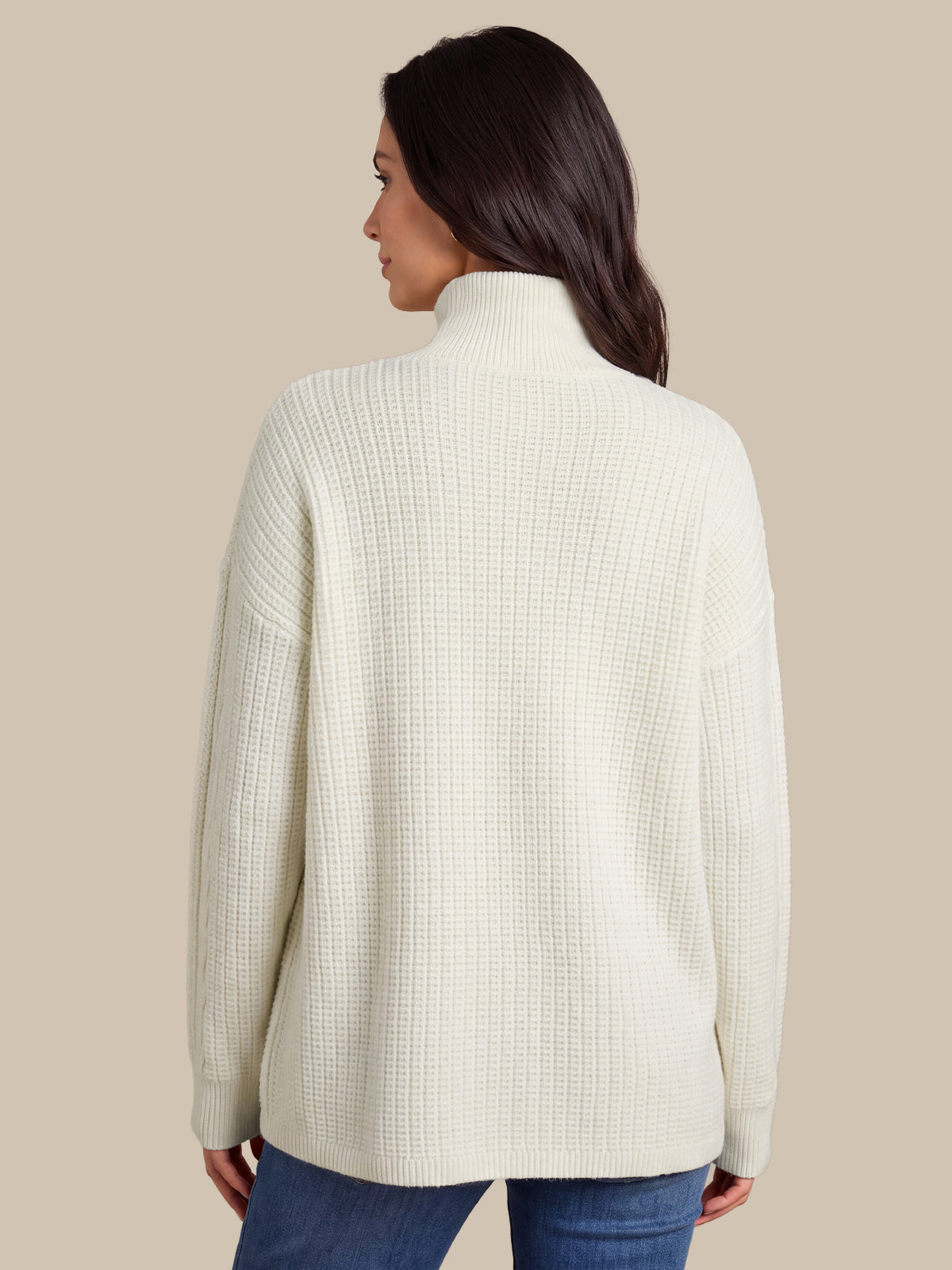 A woman shown from the back wearing a cream-colored, textured waffle-knit mock neck sweater and blue jeans, set against a solid beige background.