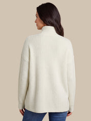 A woman shown from the back wearing a cream-colored, textured waffle-knit mock neck sweater and blue jeans, set against a solid beige background.