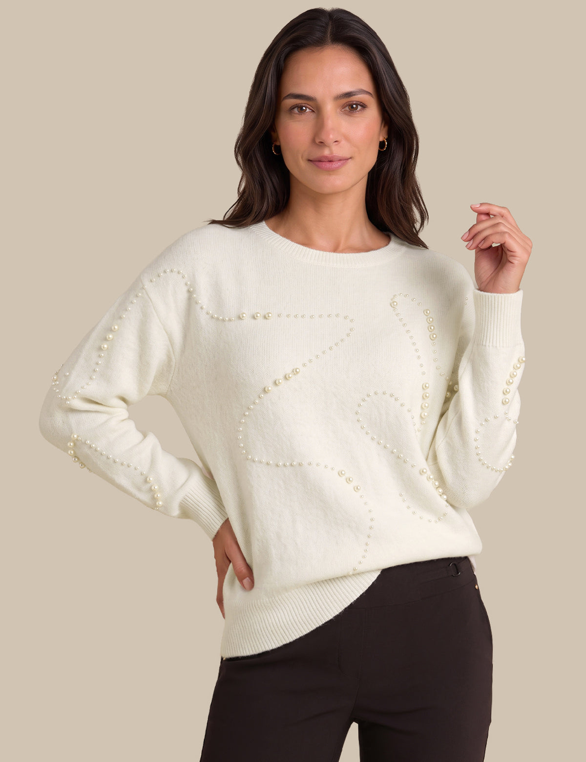 A woman modeling a cream-colored knit sweater with decorative beadwork details, paired with black pants, against a solid beige background.