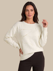 A woman modeling a cream-colored knit sweater with decorative beadwork details, paired with black pants, against a solid beige background.