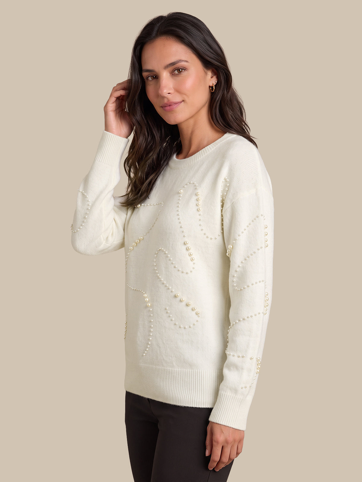 A woman modeling a cream-colored knit sweater with textured wavy patterns and pearl-like bead details, paired with dark pants, against a solid beige background.