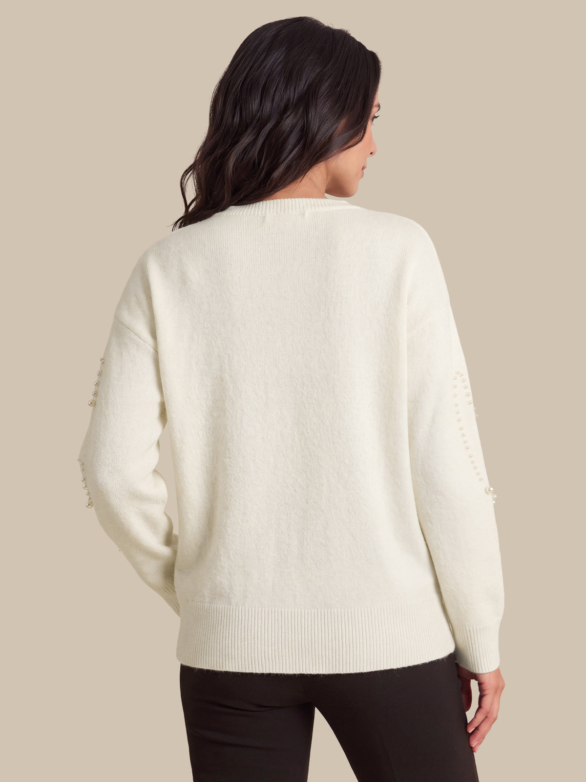 A woman viewed from the back wearing a cream-colored knit sweater with decorative bead details on the sleeves, paired with black pants, against a solid beige background.