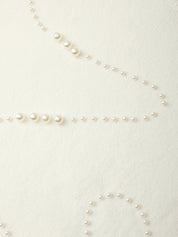 A close-up of a delicate strand of small white faux pearls laid out on a warm off-white textured fabric background.