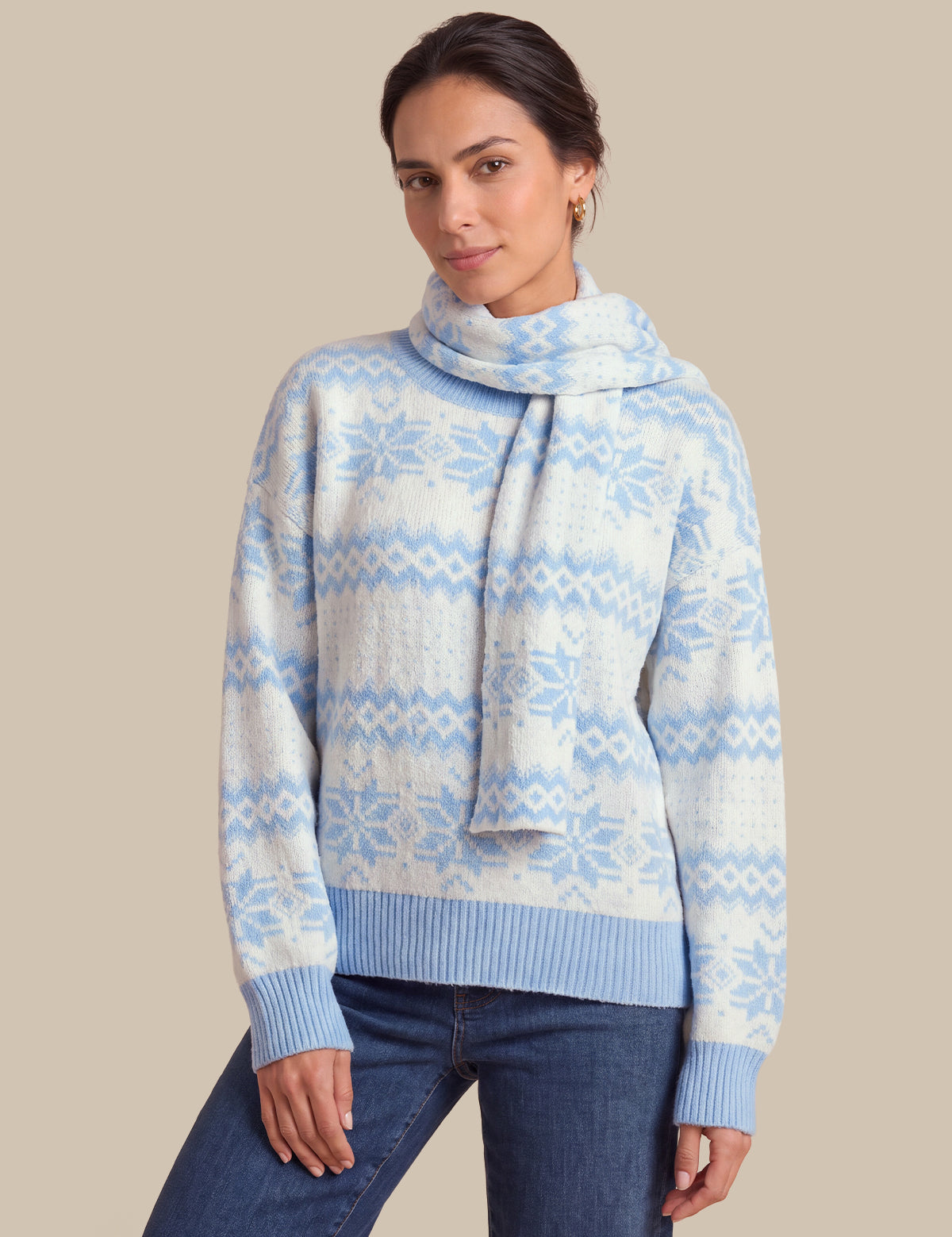 A woman wearing a light blue and white patterned knit sweater with a matching knit scarf, paired with dark blue jeans, against a solid beige background.