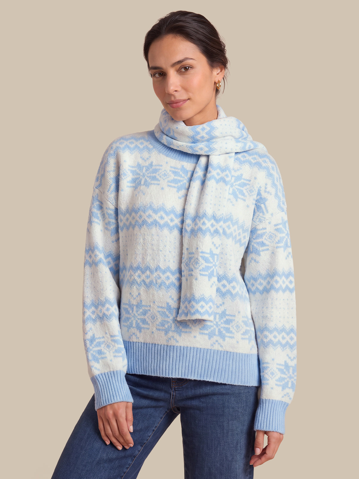 A woman wearing a light blue and white patterned knit sweater with a matching knit scarf, paired with dark blue jeans, against a solid beige background.