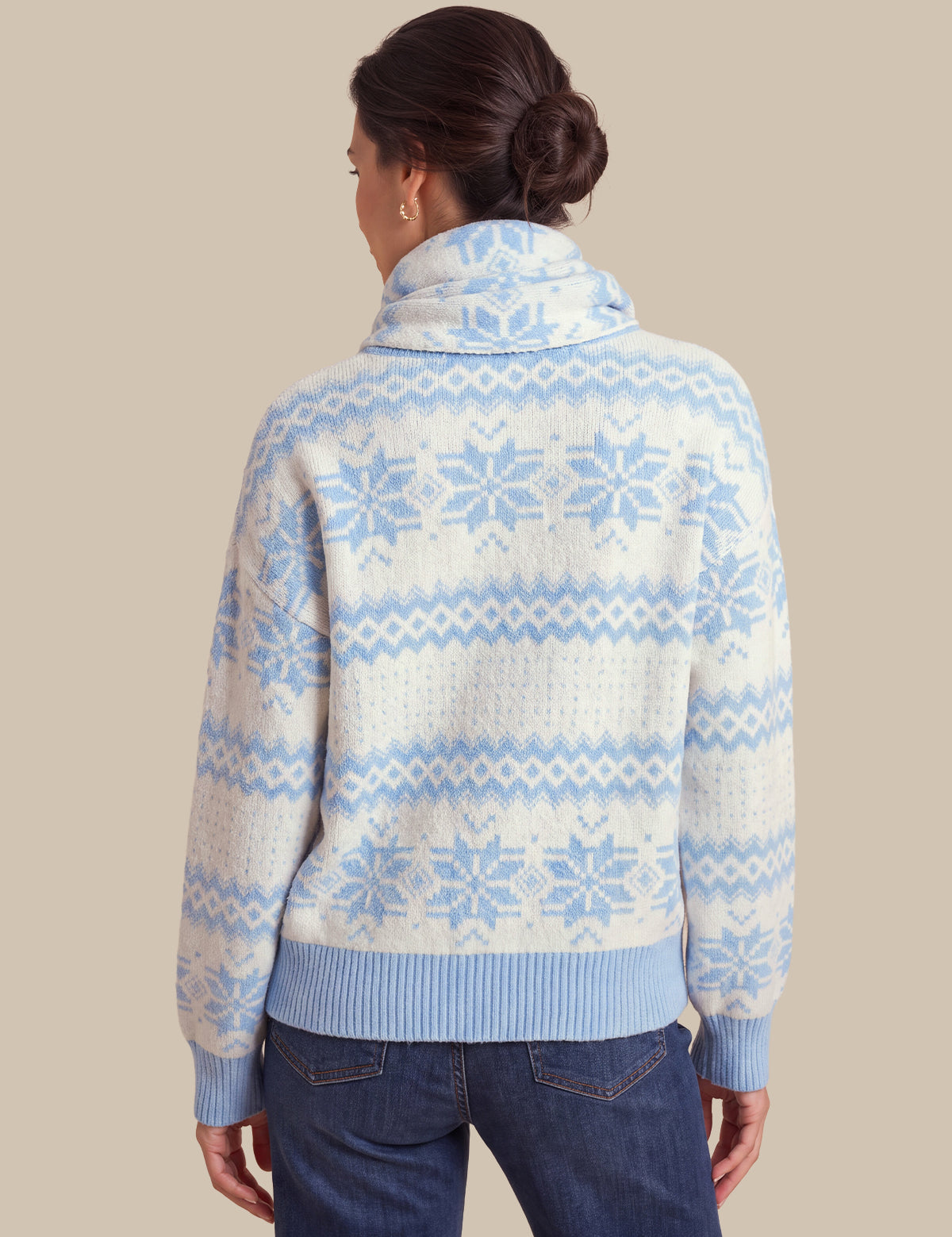 A woman shown from the back wearing a cream knit sweater with a light blue Fair Isle snowflake pattern and ribbed collar, cuffs, and hem, paired with dark blue jeans against a solid beige background.