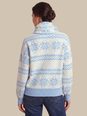 A woman shown from the back wearing a cream knit sweater with a light blue Fair Isle snowflake pattern and ribbed collar, cuffs, and hem, paired with dark blue jeans against a solid beige background.