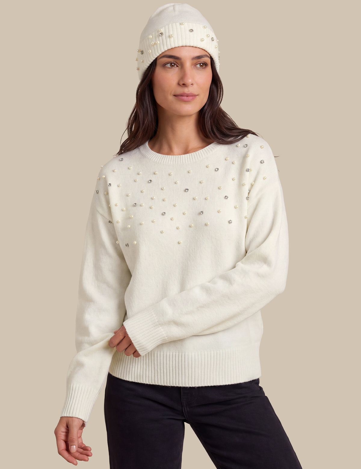 A woman wearing a cream-colored knit sweater with small silver rhinestone embellishments, paired with black pants and a matching beanie, set against a solid beige background.