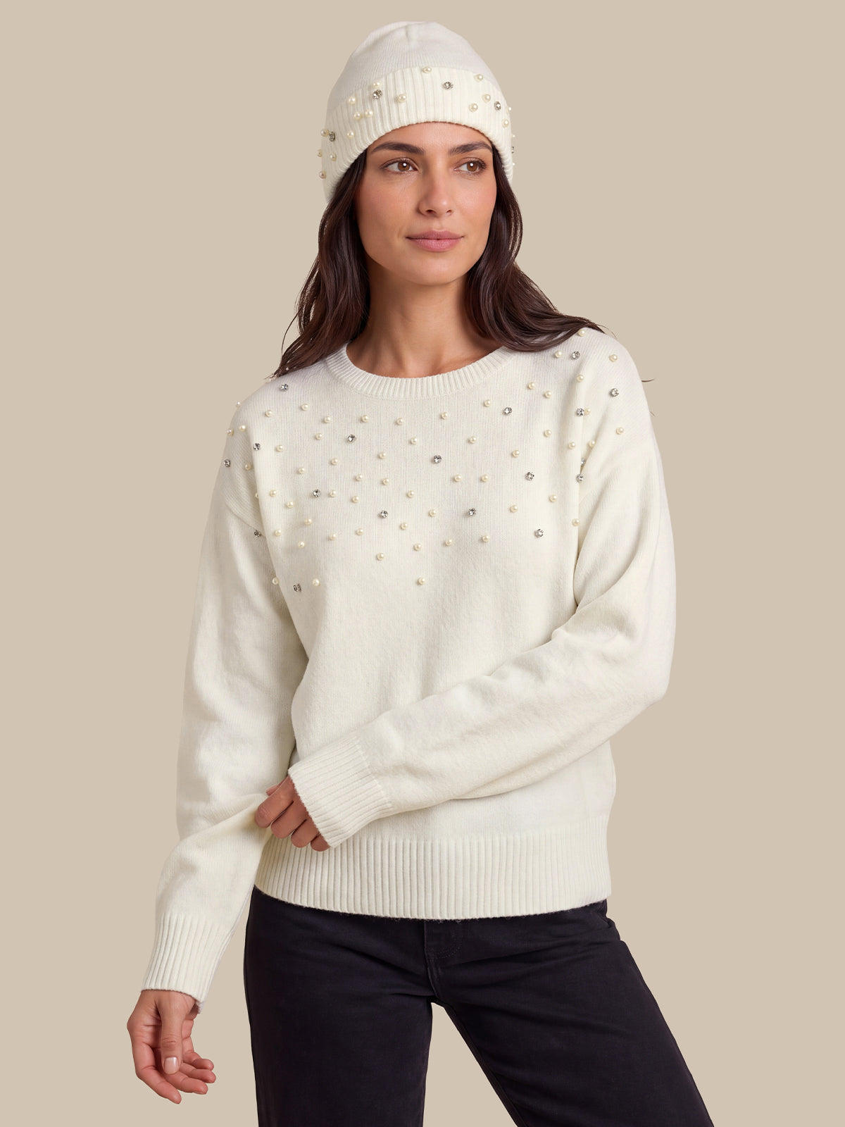 A woman wearing a cream-colored knit sweater with small silver rhinestone embellishments, paired with black pants and a matching beanie, set against a solid beige background.