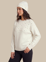 A woman wearing a cream knit sweater with small metallic embellishments across the chest, paired with a matching cream beanie and dark pants, against a solid beige background.