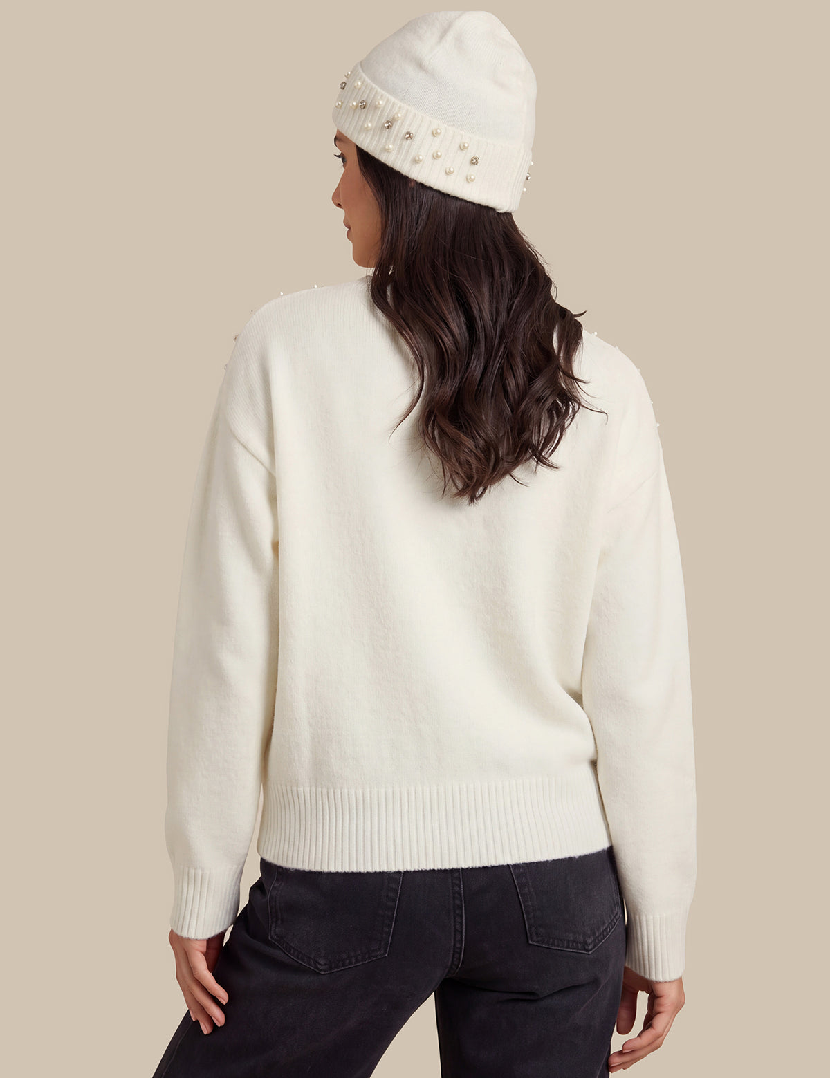 A woman shown from the back wearing an off-white knit sweater, a matching off-white beanie with small metallic embellishments, and dark jeans, against a solid beige background.