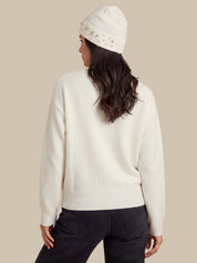 A woman shown from the back wearing an off-white knit sweater, a matching off-white beanie with small metallic embellishments, and dark jeans, against a solid beige background.
