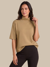 A woman modeling a short-sleeve, mock-neck knit top in camel beige, paired with black pants, against a solid beige background.