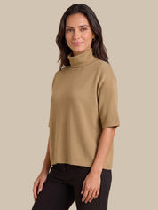 A woman modeling a short-sleeve, high-neck turtleneck top in camel beige, paired with black pants, against a solid beige studio background.