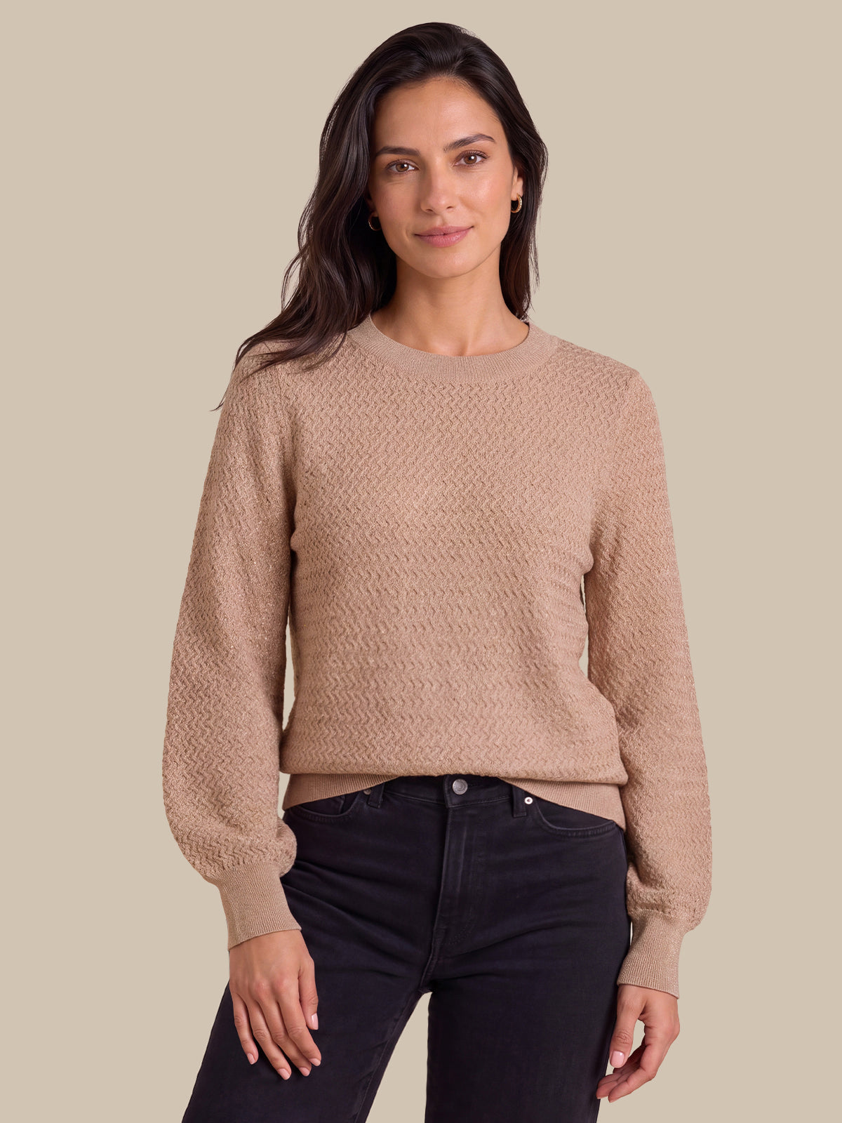 A woman wearing a textured, long-sleeve crewneck sweater in a warm beige/tan color, paired with dark jeans, against a solid beige background.
