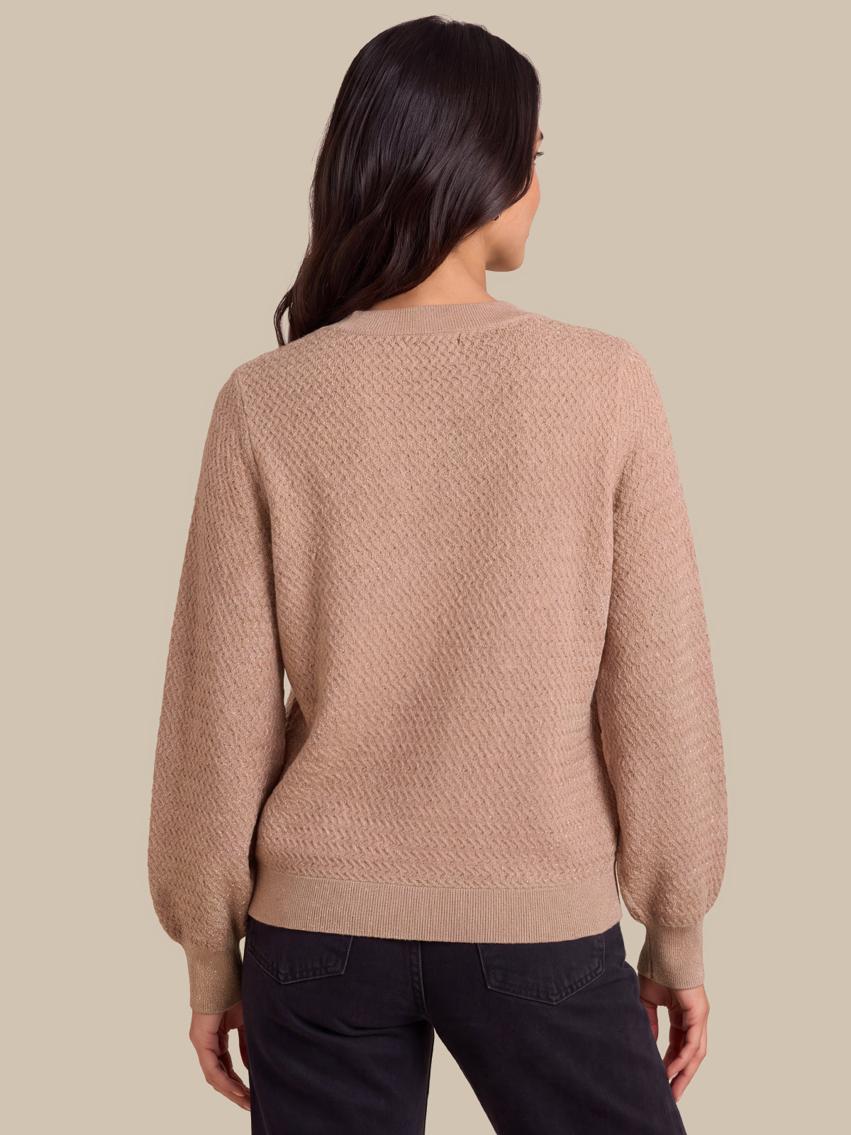 A woman viewed from the back wearing a textured knit sweater in a dusty rose-beige color, paired with dark jeans, against a solid beige background.