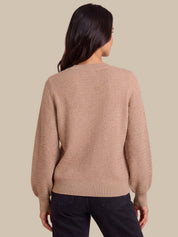 A woman viewed from the back wearing a textured knit sweater in a dusty rose-beige color, paired with dark jeans, against a solid beige background.