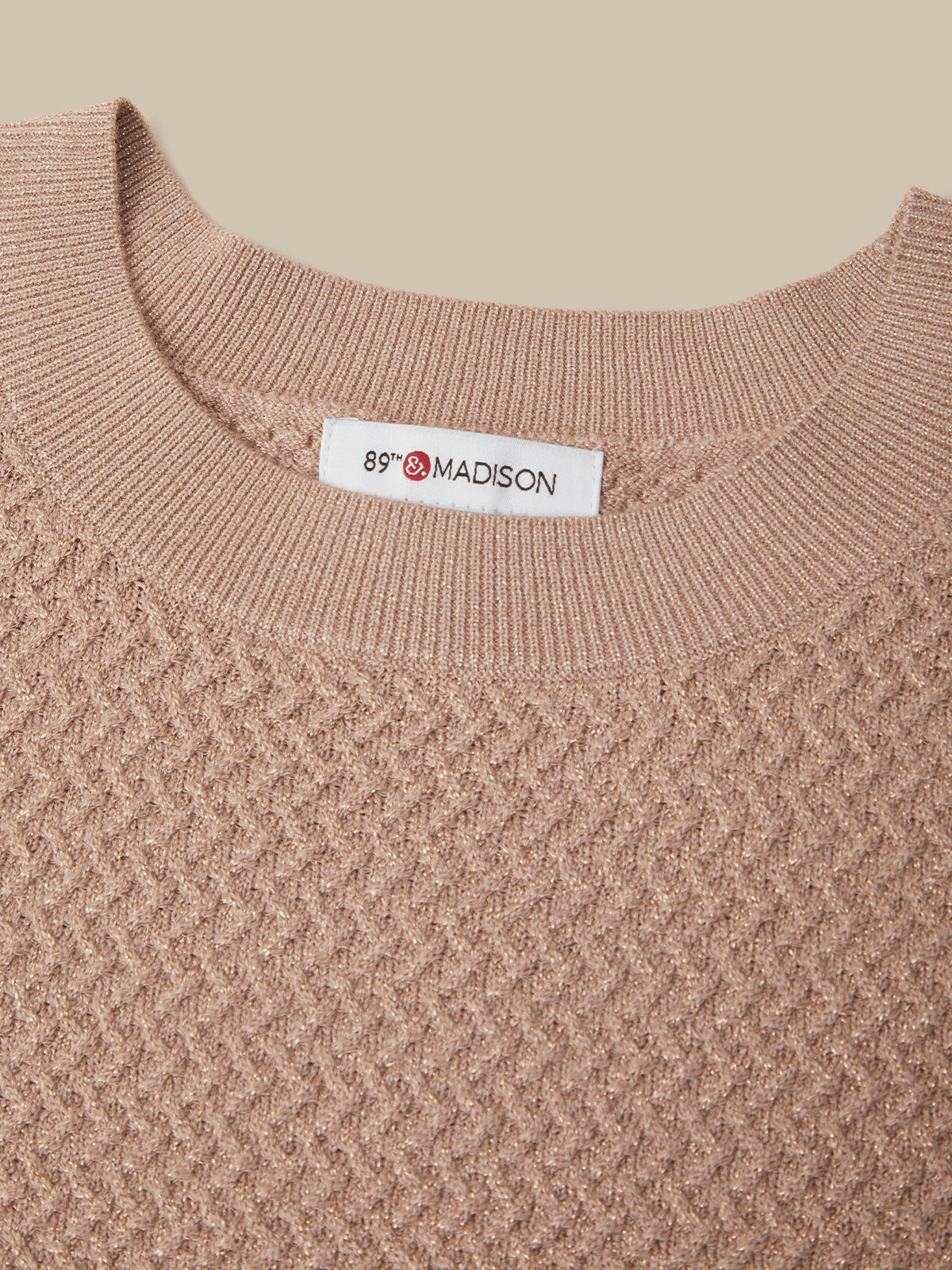 Close-up of a textured knit crewneck sweater in a warm taupe-beige color, featuring a white neck label with the brand name '89 MADISON'.