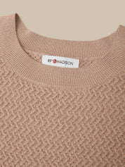 Close-up of a textured knit crewneck sweater in a warm taupe-beige color, featuring a white neck label with the brand name '89 MADISON'.