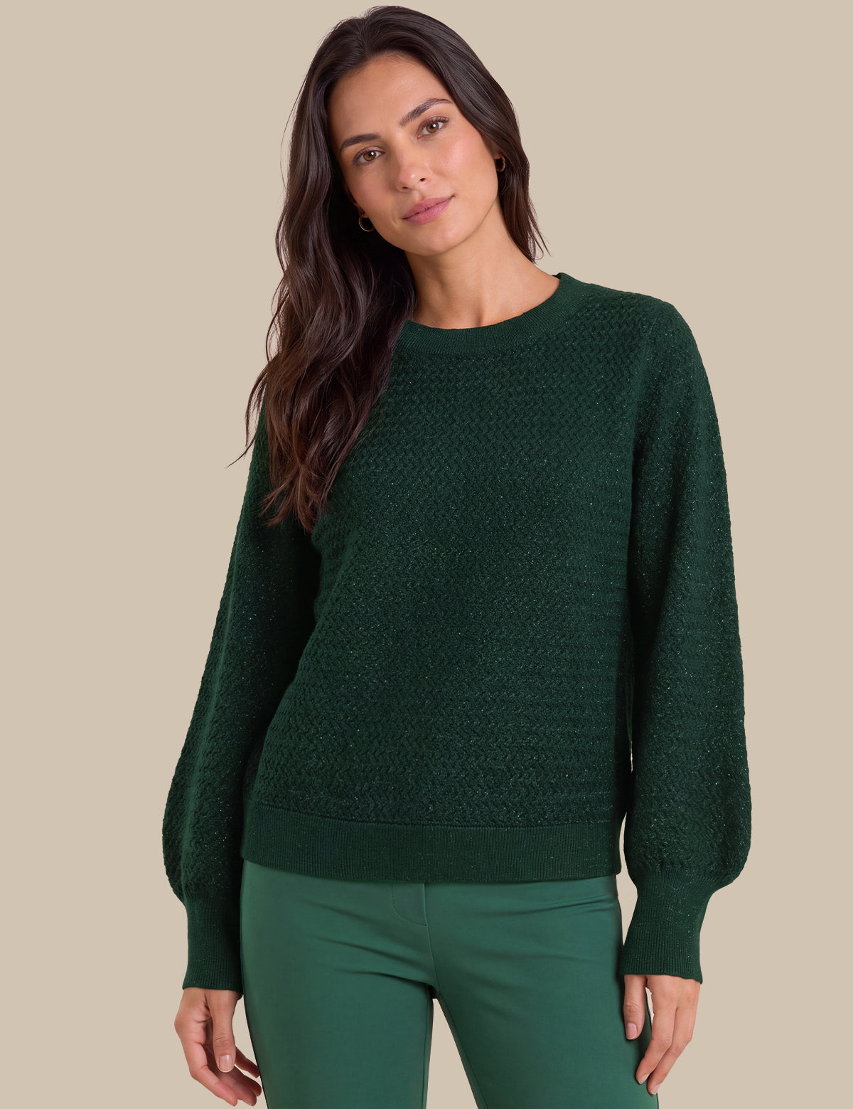 A woman wearing a dark green textured knit sweater with balloon sleeves and matching green pants, posed against a solid beige background.
