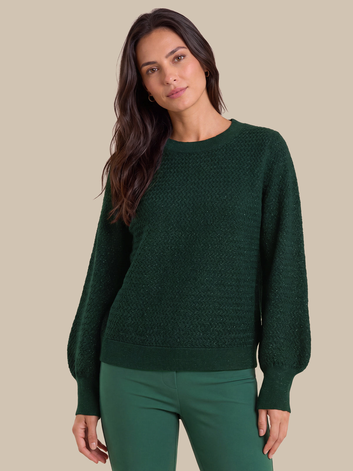 A woman wearing a dark green textured knit sweater with balloon sleeves and matching green pants, posed against a solid beige background.