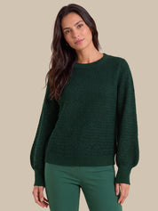 A woman wearing a dark green textured knit sweater with balloon sleeves and matching green pants, posed against a solid beige background.