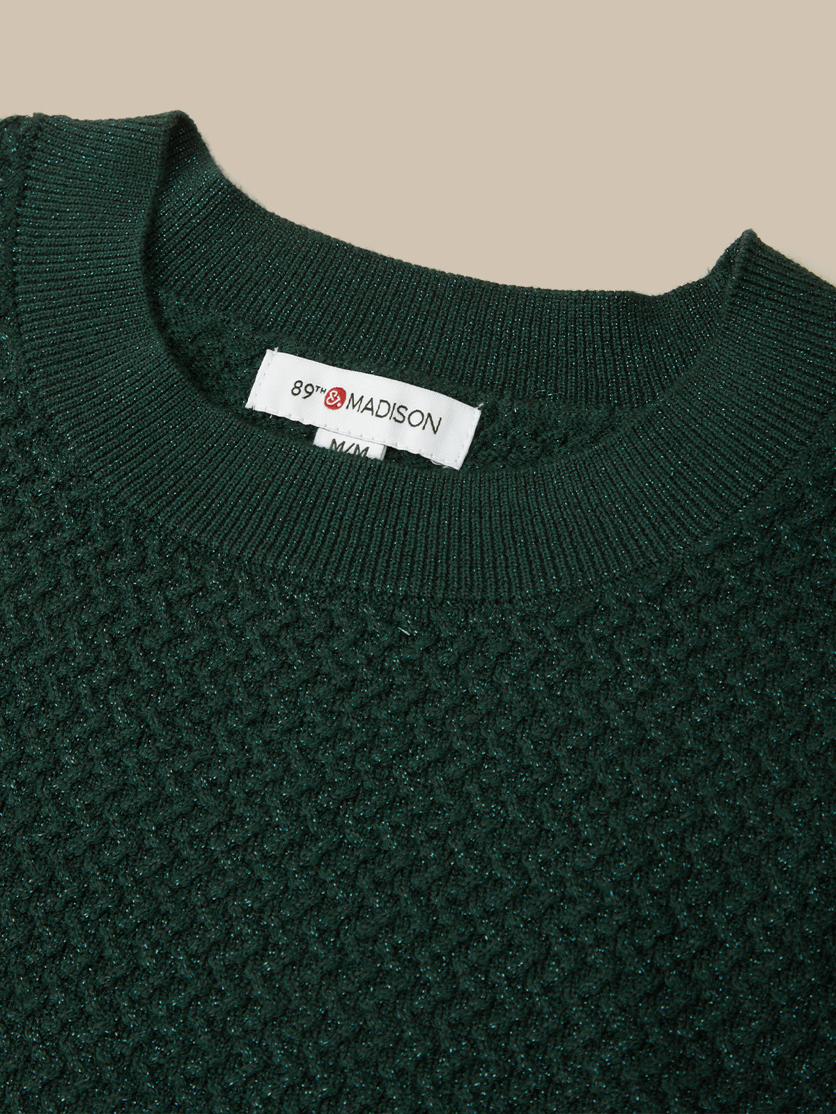 Close-up of a dark forest green knit sweater with a textured waffle pattern, featuring a white neck label that reads '89° MADISON'.