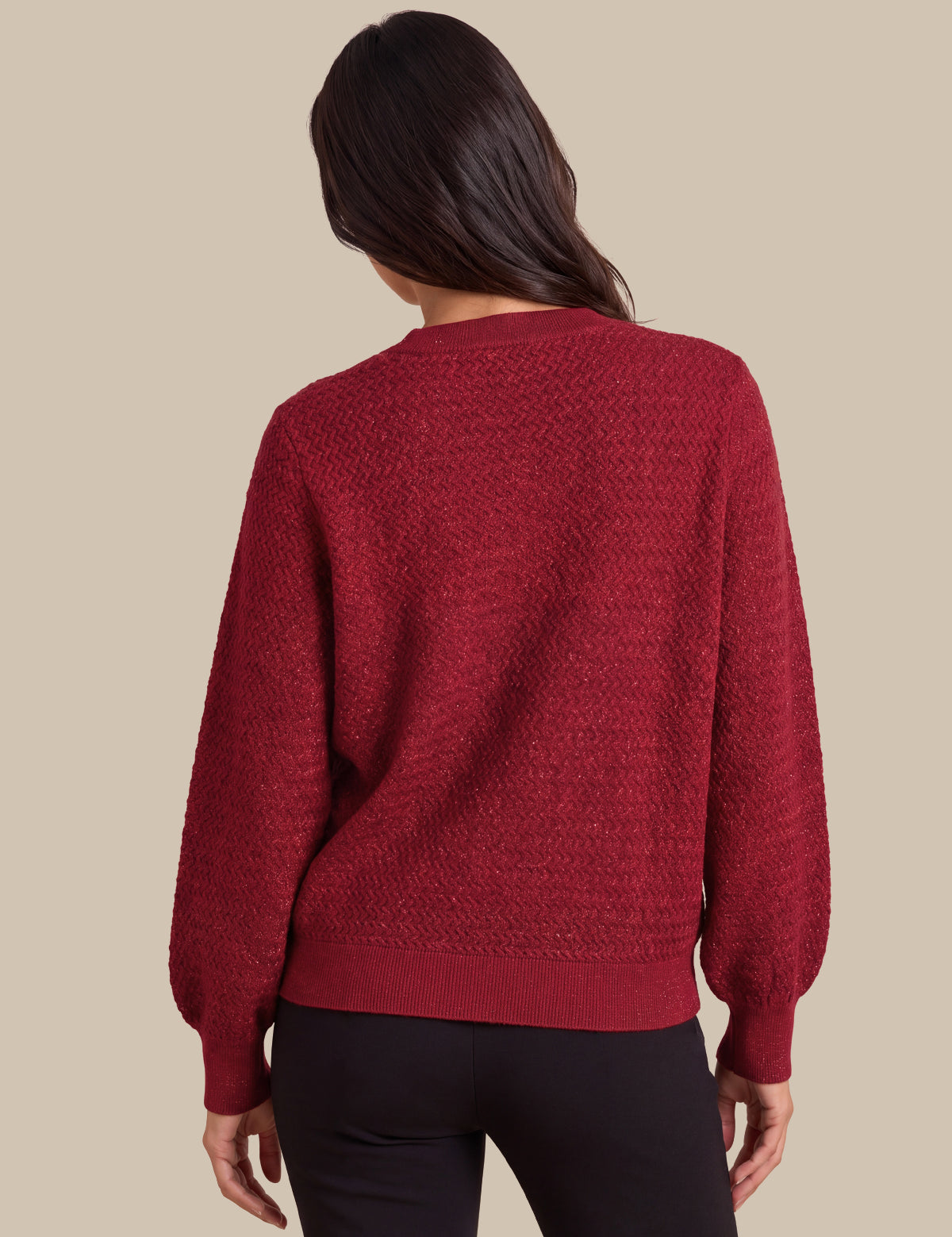 A woman viewed from the back wearing a textured, deep red knit sweater with balloon sleeves and black pants, set against a solid beige background.