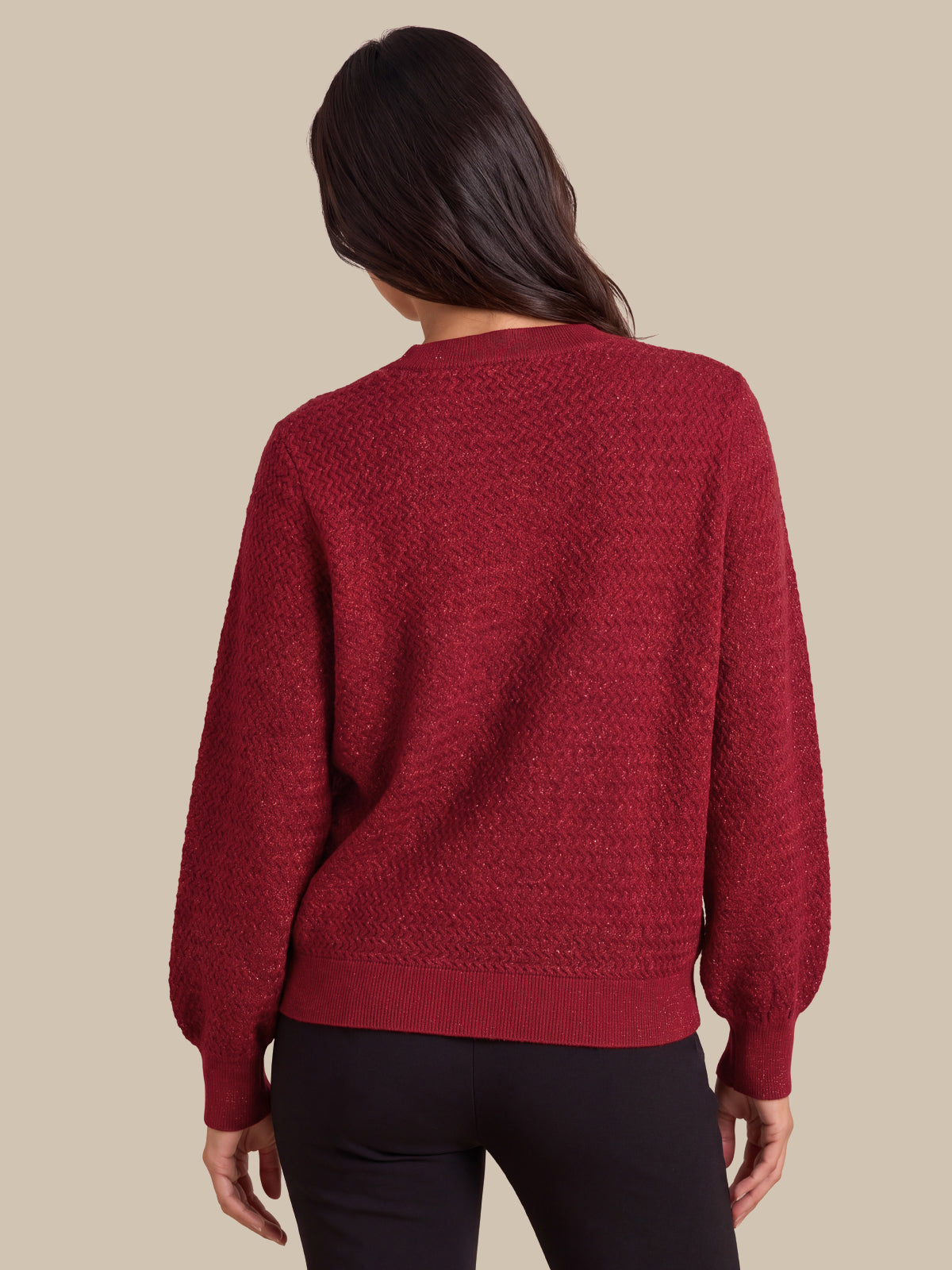A woman viewed from the back wearing a textured, deep red knit sweater with balloon sleeves and black pants, set against a solid beige background.