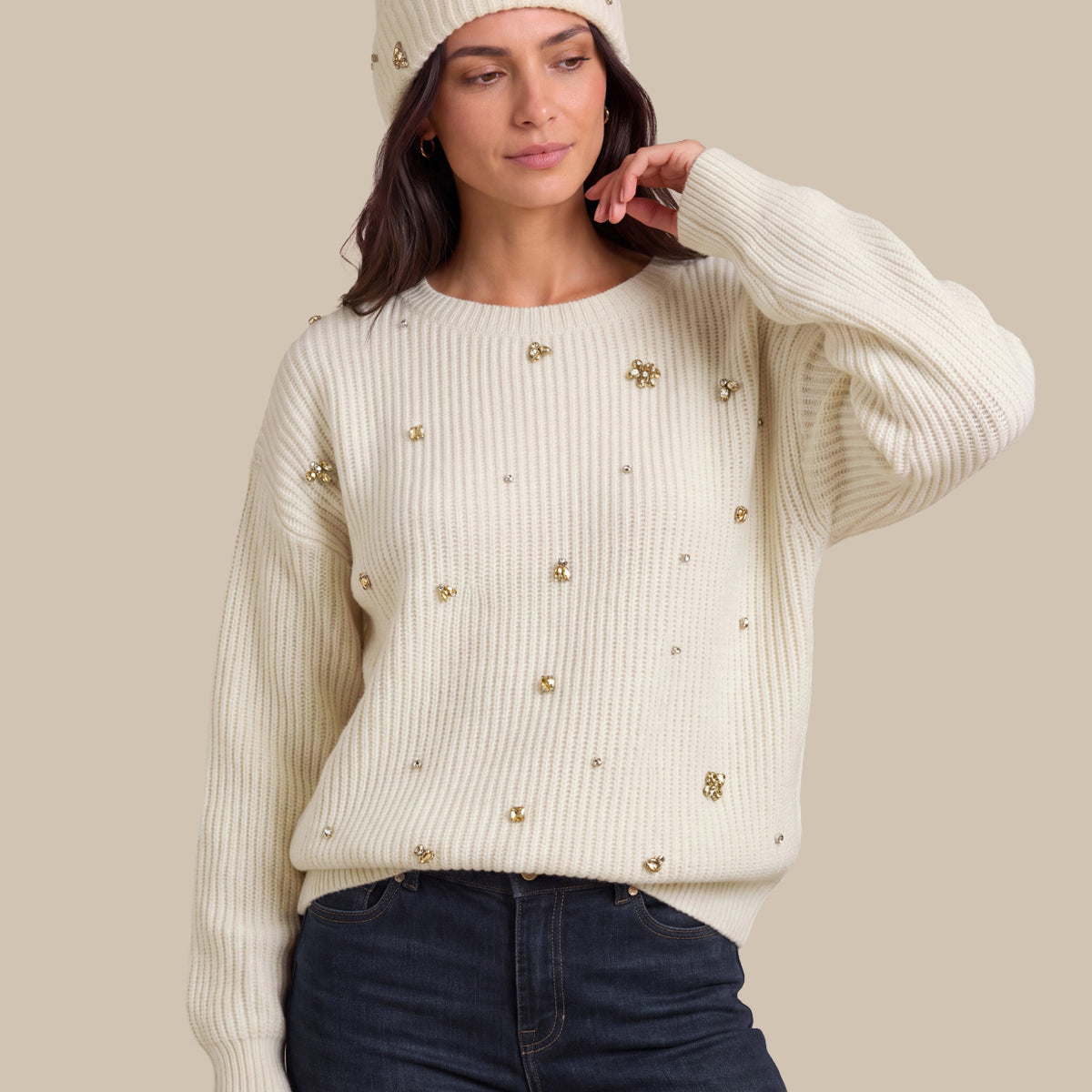A woman wearing a cream ribbed knit sweater with gold floral embellishments, a matching cream beanie with a pom-pom, and dark blue jeans, posed against a solid beige background.