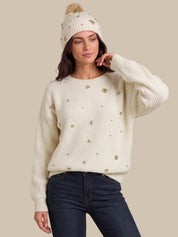 A woman wearing a cream ribbed knit sweater with gold floral embellishments, a matching cream beanie with a pom-pom, and dark blue jeans, posed against a solid beige background.