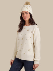 A woman wearing a cream ribbed knit sweater with gold embroidered details, a matching cream knit beanie with a pom-pom, and dark blue jeans, set against a solid beige background.