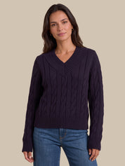 A woman wearing a dark purple cable-knit V-neck sweater and blue jeans, standing against a solid beige background.