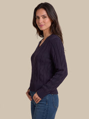 A woman wearing a dark purple cable-knit V-neck sweater and blue jeans, shown from a side profile against a solid beige background.