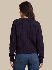 A woman shown from the back wearing a dark navy cable-knit sweater and blue jeans against a solid beige background.