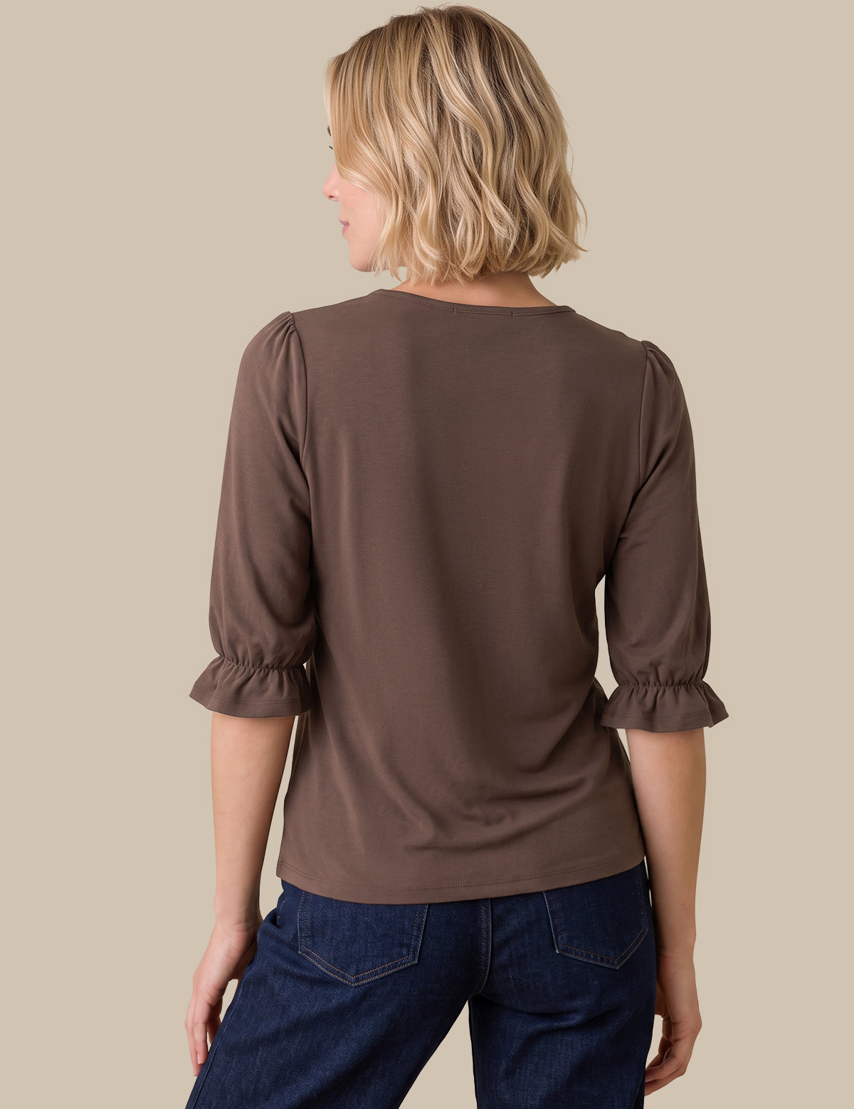 A woman shown from the back wearing a brown three-quarter sleeve top with ruffled cuffs and dark blue jeans, set against a solid beige background.